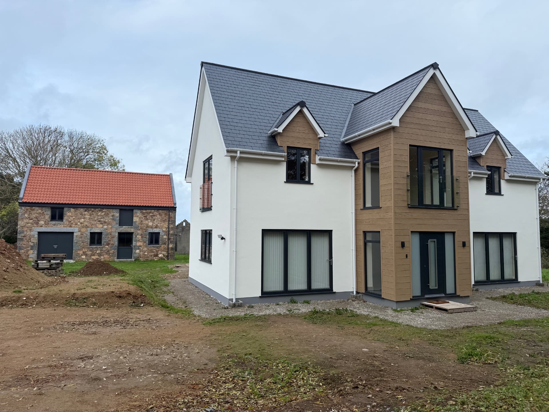 Finished newbuild home with white render, timber cladding and slate roof beside a restored stone barn